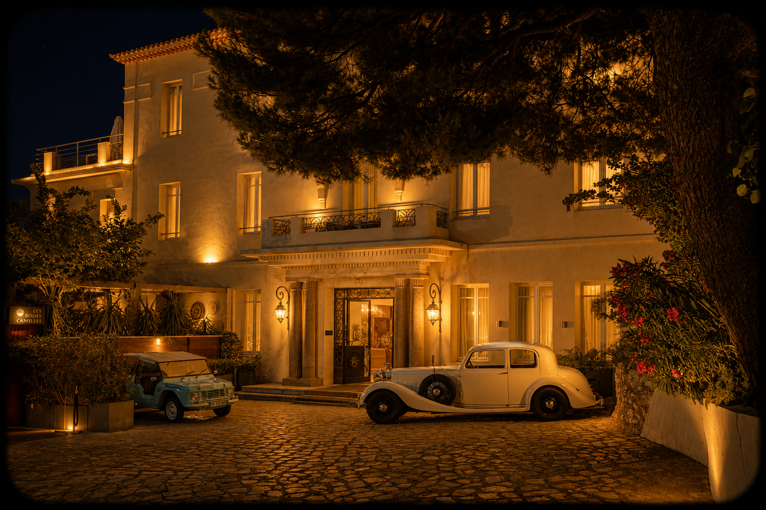 Un bâtiment élégant illuminé la nuit avec des voitures anciennes garées devant, entouré de plantes et d'arbres.