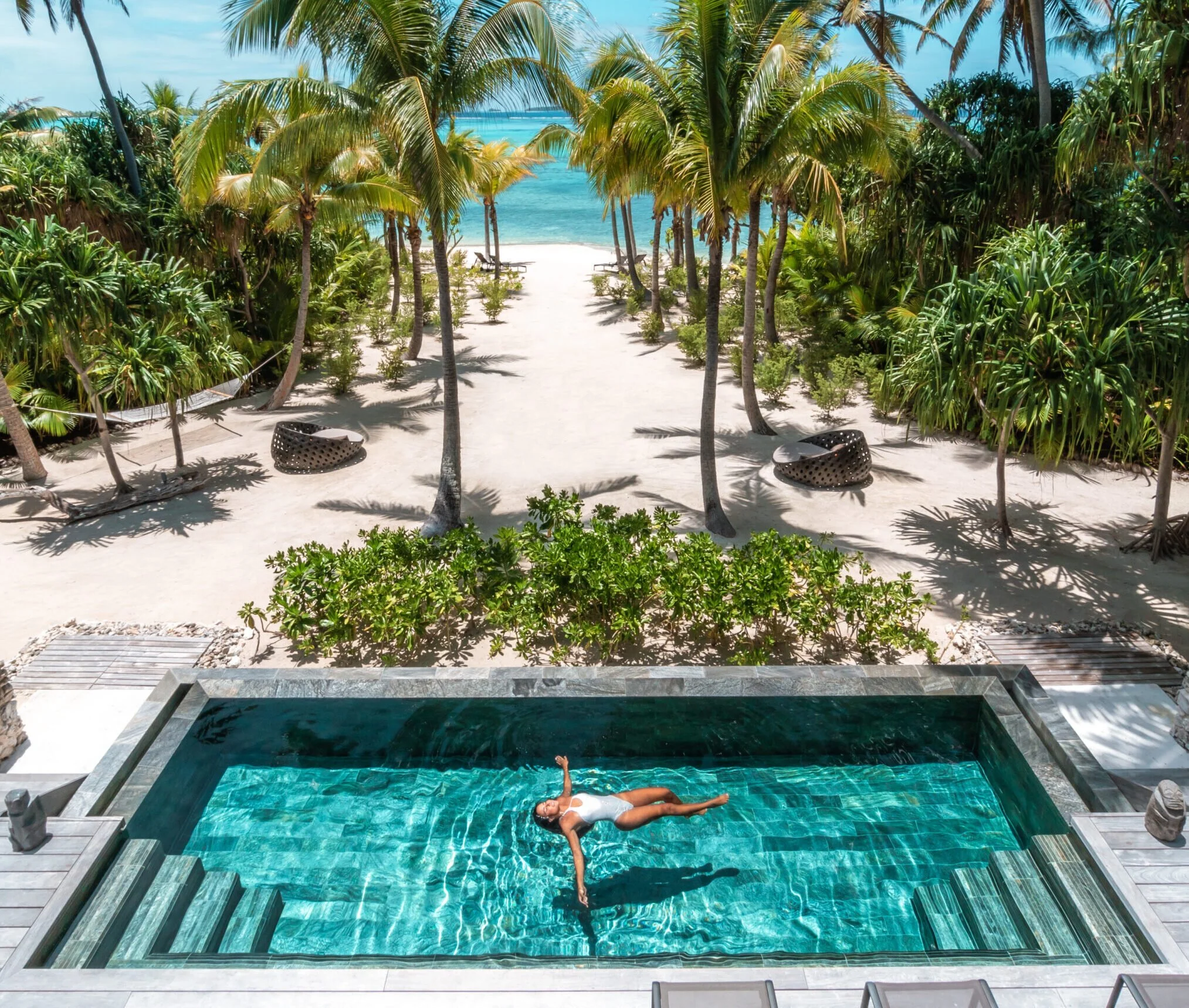 Une femme nage dans une piscine face à une plage bordée de palmiers et l'océan bleu au loin, sous un ciel ensoleillé.