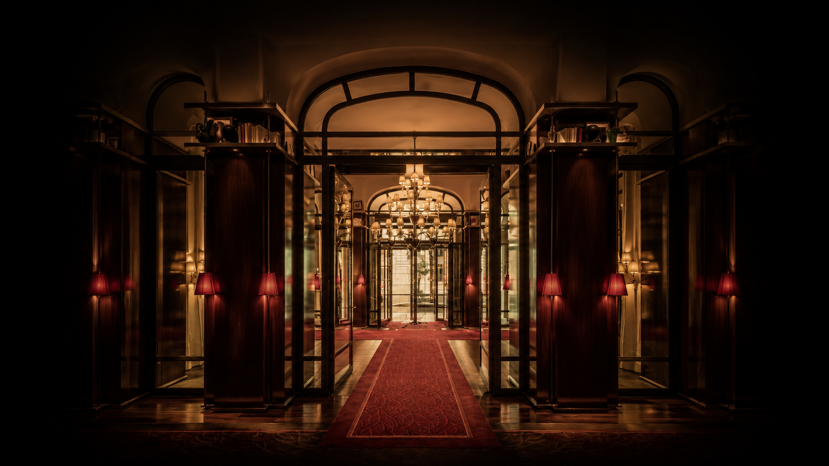 Un couloir d'entrée élégant avec un tapis rouge, des lampes à abat-jour rouge sur le mur et des chandeliers suspendus au plafond, menant à une porte vitrée.