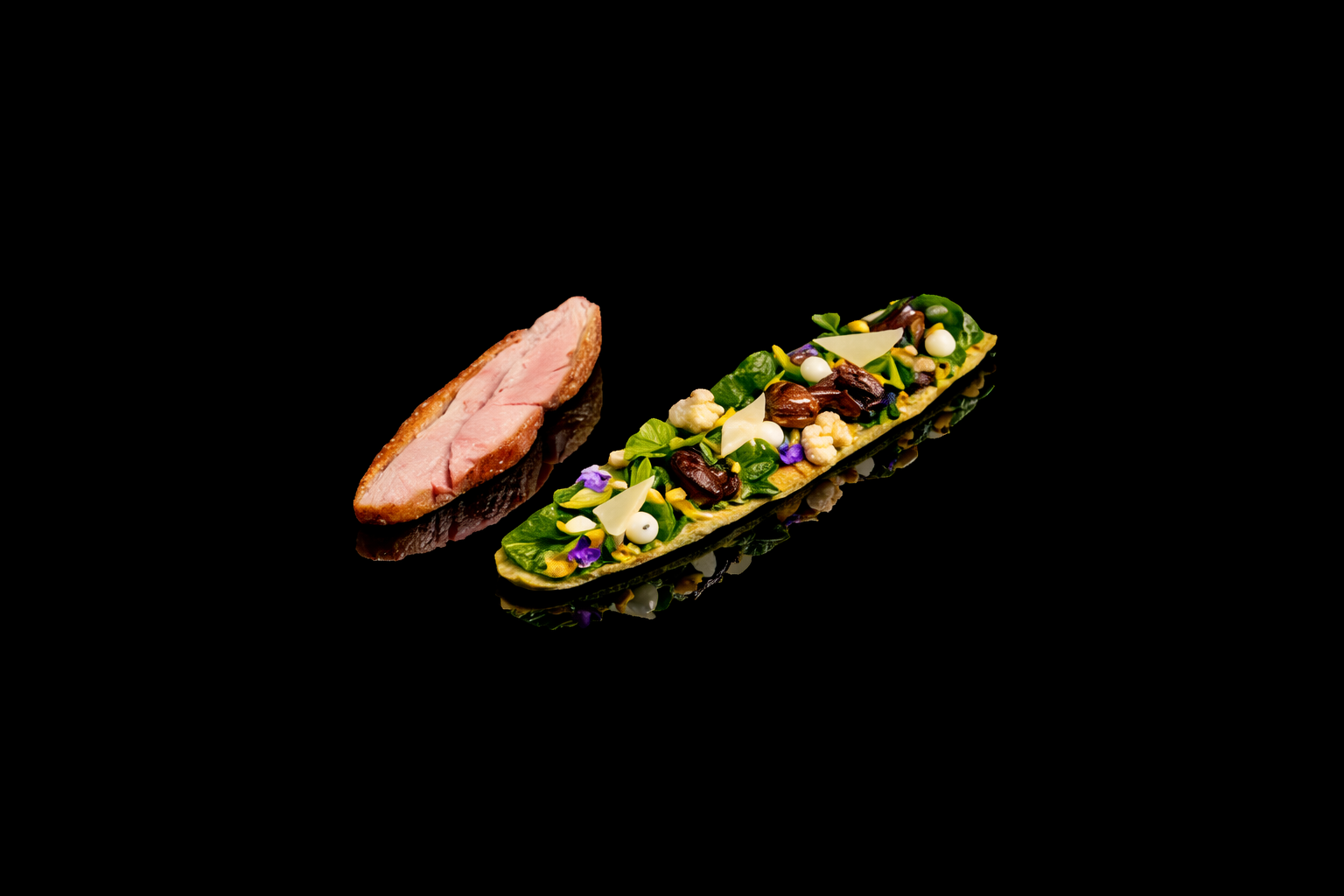 Une assiette de viande cuite en tranches et une bouchée végétale garnie d'herbes, de fleurs comestibles, de noix et d'autres garnitures sur fond noir.