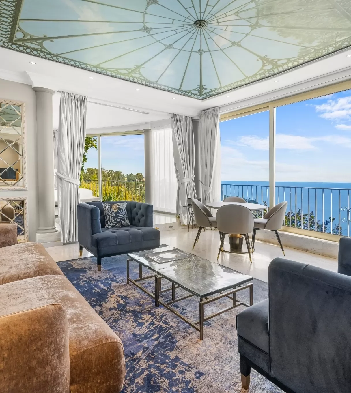 Salon avec canapé beige, fauteuil noir, table basse en verre, grandes fenêtres avec vue sur la mer et la nature, murs blancs, plafond décoratif avec motif d'horloge, rideaux blancs, mobilier moderne.