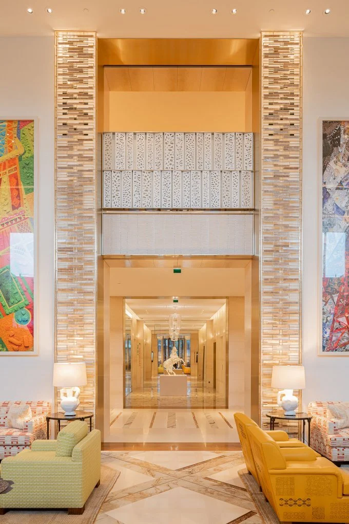 Un grand hall d'hôtel ou d'immeuble avec des canapés jaunes et verts, des lampes à côté, des œuvres d'art colorées sur les murs, et une arche menant à un couloir avec une sculpture d'un animal en son centre.