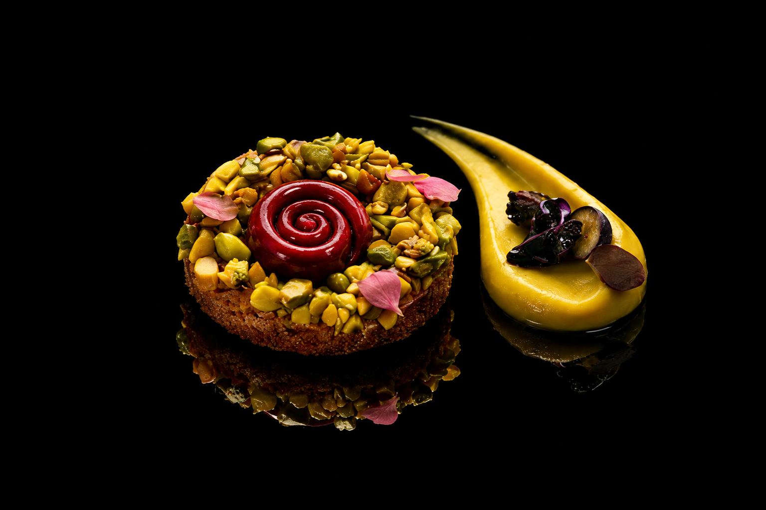 Un gâteau rond décoré de pistaches, de fleurs en pétales roses, avec un glaçage rouge en spirale au centre, et une sauce jaune avec des garnitures de petits fruits et fleurs en pétales, sur un fond noir.