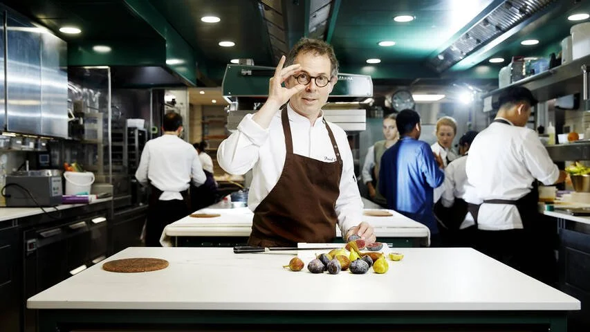 Chef professionnel dans une cuisine de restaurant, portant un tablier marron et une chemise blanche, se tenant derrière un plan de travail blanc avec des figues et un gâteau rond, tout en ajustant ses lunettes et regardant vers la caméra.