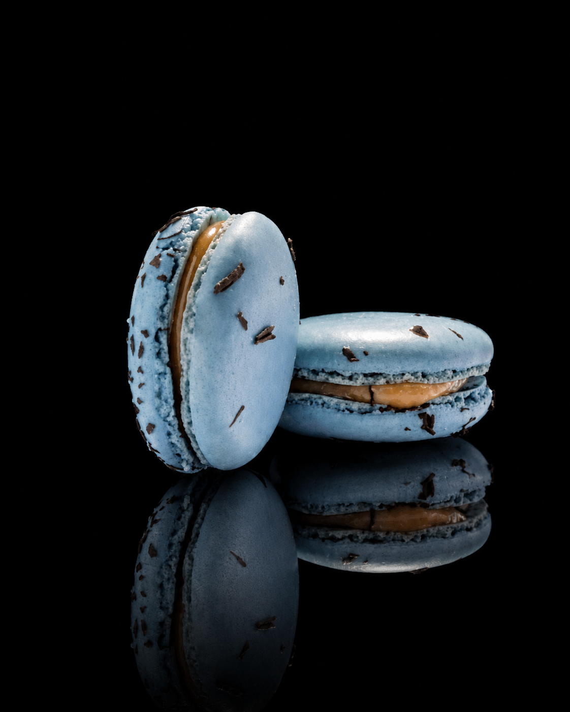 Deux macarons bleus avec des éclats de chocolat, reflétés sur une surface noire.