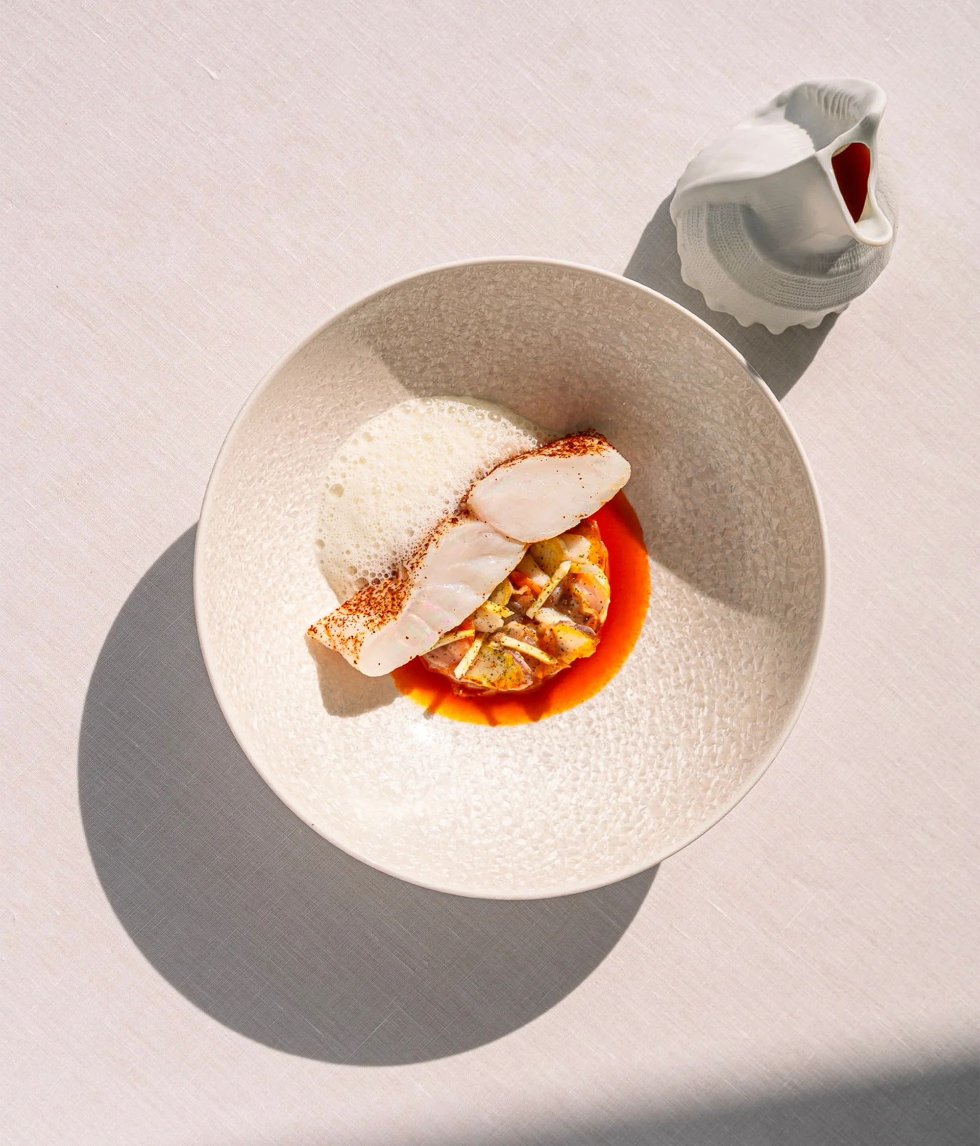 Une assiette avec un plat sophistiqué, comprenant une portion de poisson blanc grillé, un mélange de fruits de mer ou légumes, une sauce orange, et une mousse blanche en garniture. À côté, une cruche en céramique blanche avec un extérieur texturé, pour verser une sauce ou un liquide.