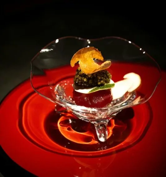 Un dessert sophistiqué dans un bol en verre, avec une garniture de zeste d'orange, une boule de sorbet, et d'autres éléments décoratifs sur un plateau rouge
