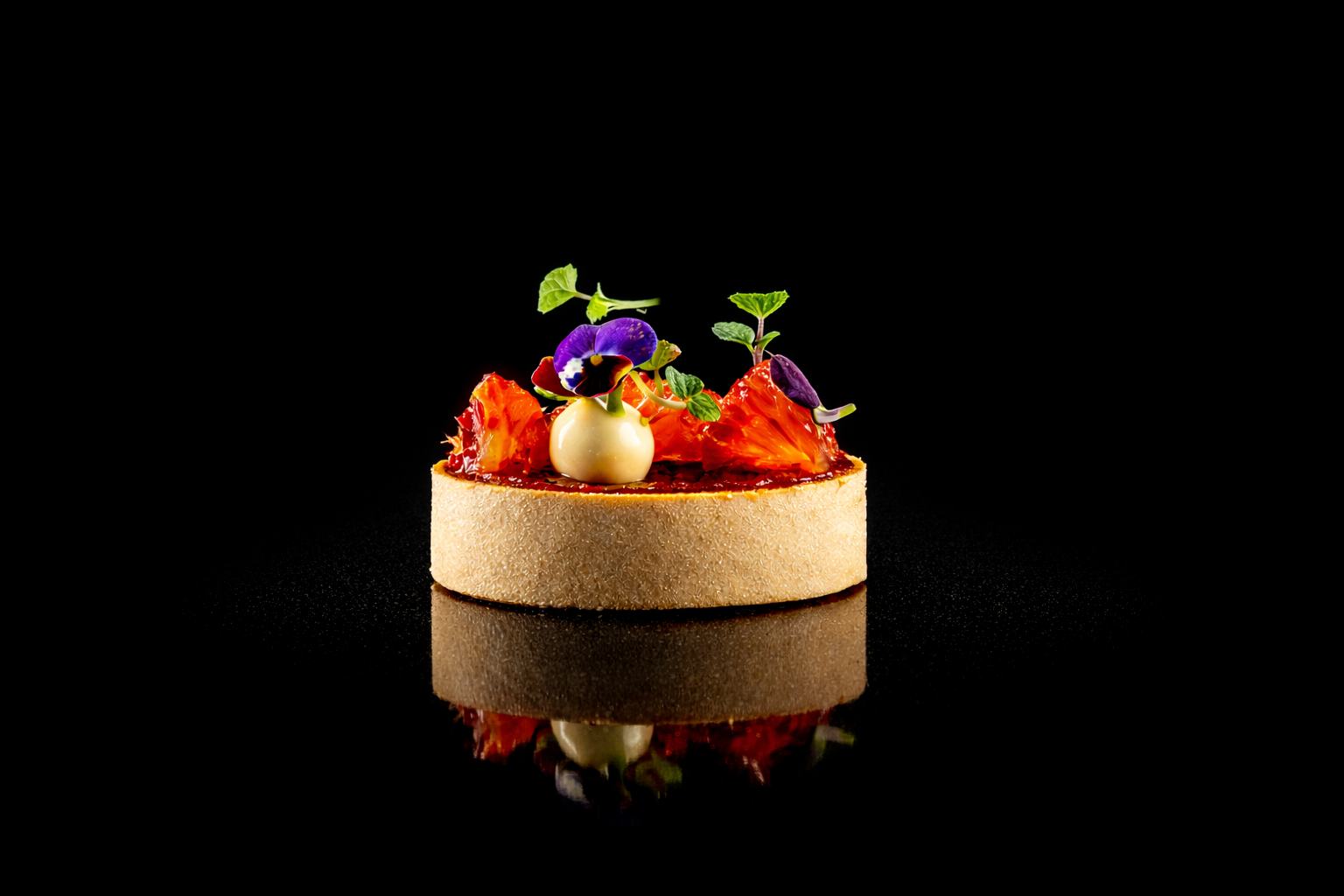 Un dessert gastronomique présenté sur une plaque noire, avec une base ronde beige, garnie de morceaux de fruits, de fleurs comestibles et d'une petite boule blanche.
