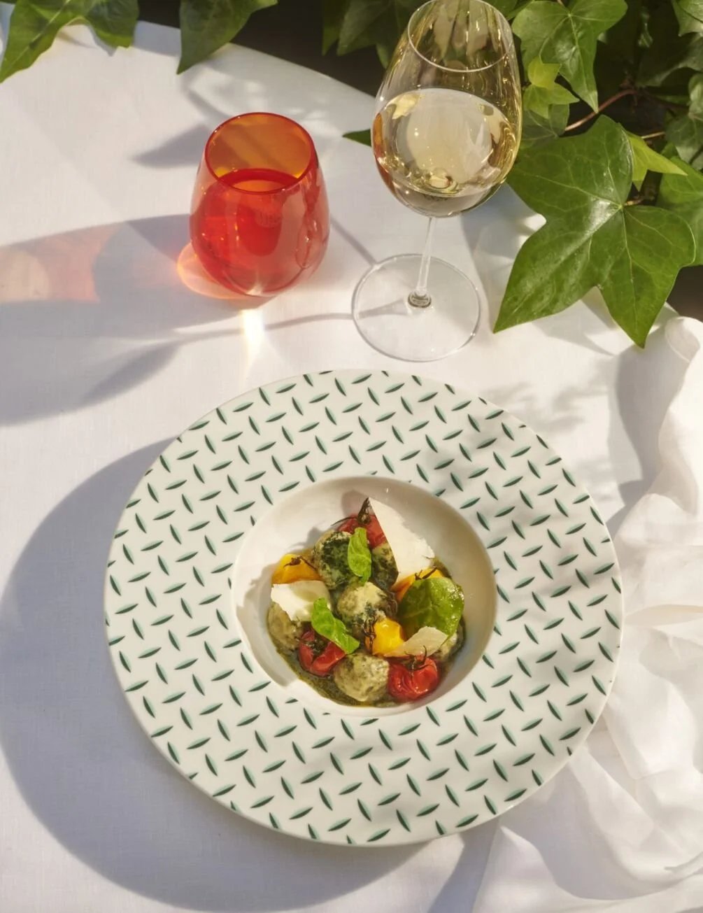 Une assiette de boulettes de viande avec des légumes verts, des tomates cerises et de la mozzarella sur une table blanche avec un verre de vin blanc et un verre de boisson orange, entourés de feuilles vertes.