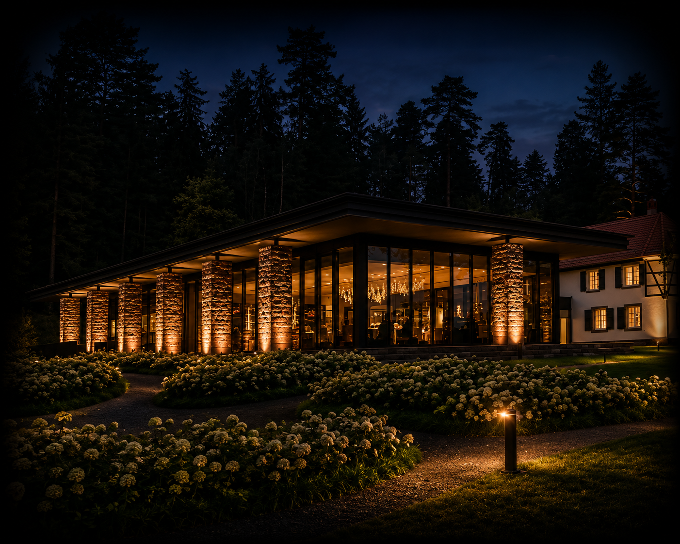 Une maison moderne en verre illuminée la nuit, entourée de jardins de fleurs et de grands arbres forestiers.