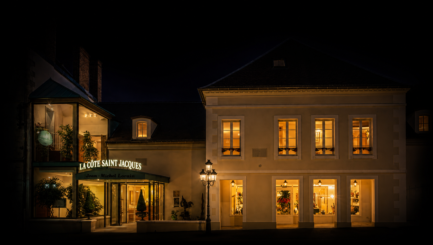 Bâtiment lumineux la nuit avec une enseigne indiquant 'La Côte Saint Jacques' et des fenêtres allumées, décorée avec des plantes et un lampadaire en devanture.