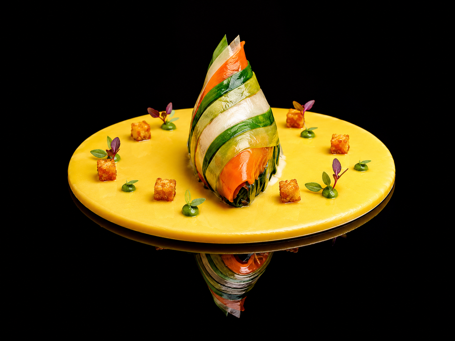 Plat de cuisine moderne avec un fromage jaune rond, décoré d'une sculpture faite de tranches de légumes colorés, entouré de petits cubes croustillants et de feuilles vertes