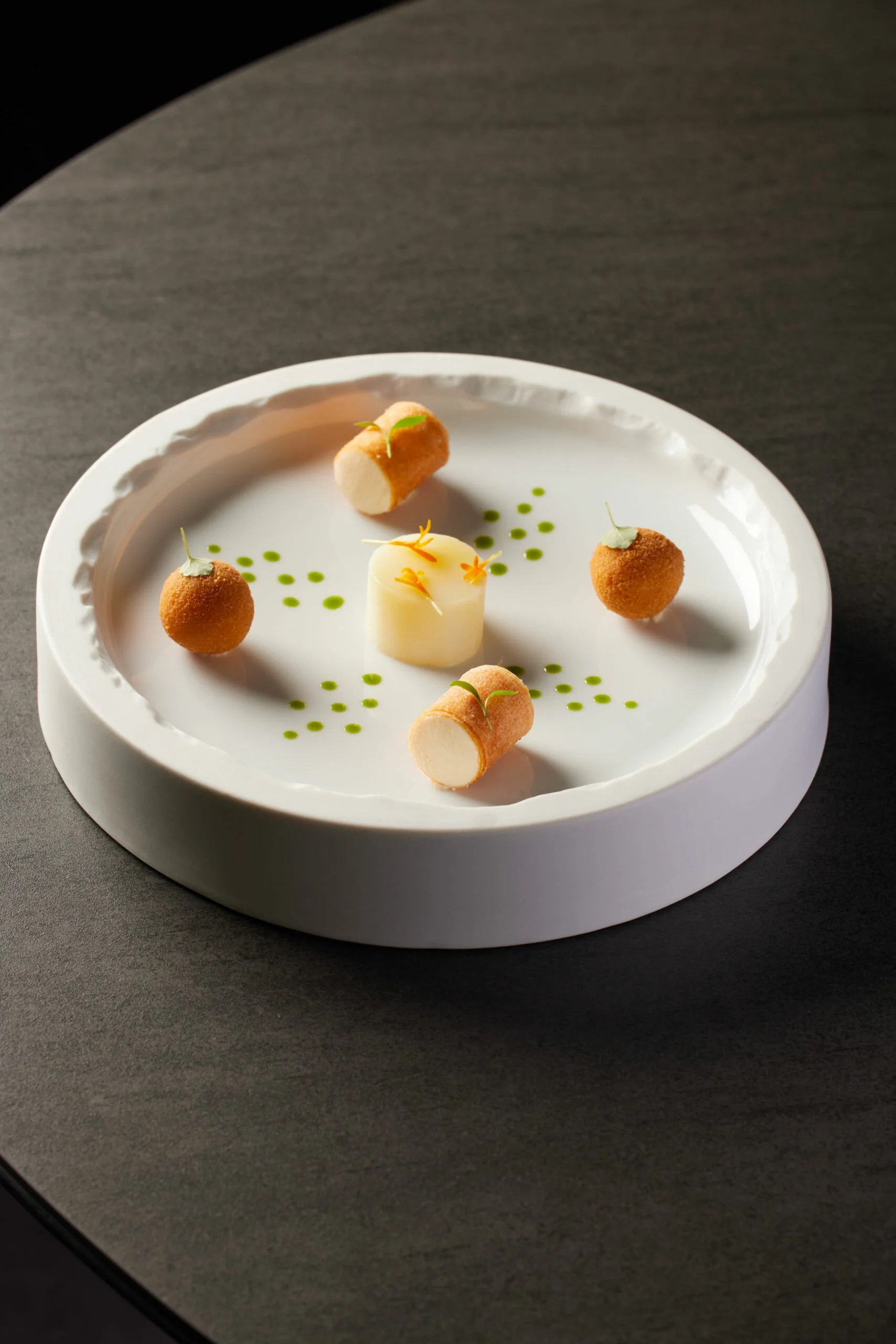 Assiette de amuse-bouches avec quatre boulettes dorées, un cylindre blanc et petites touches de vert et d'orange décoratives.