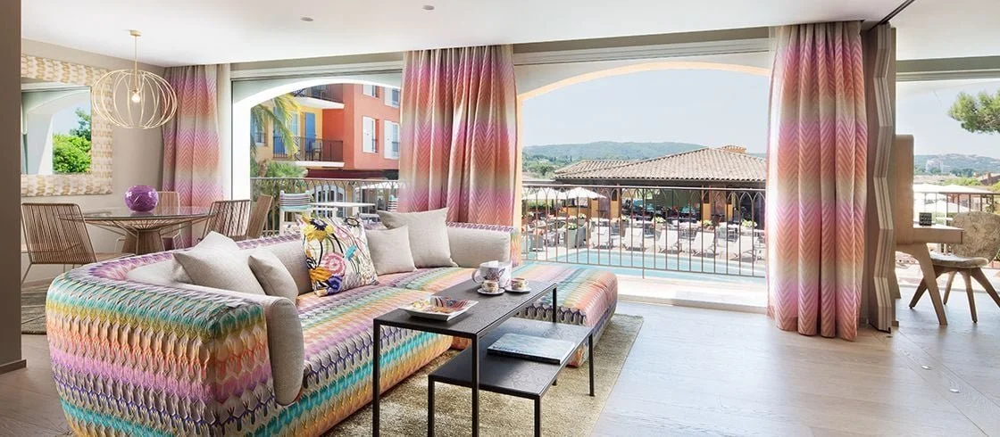 Salon lumineux avec canapé coloré, grande fenêtre avec vue, rideaux à motifs pastel, meubles modernes, terrasse avec parasol et vue sur la ville.