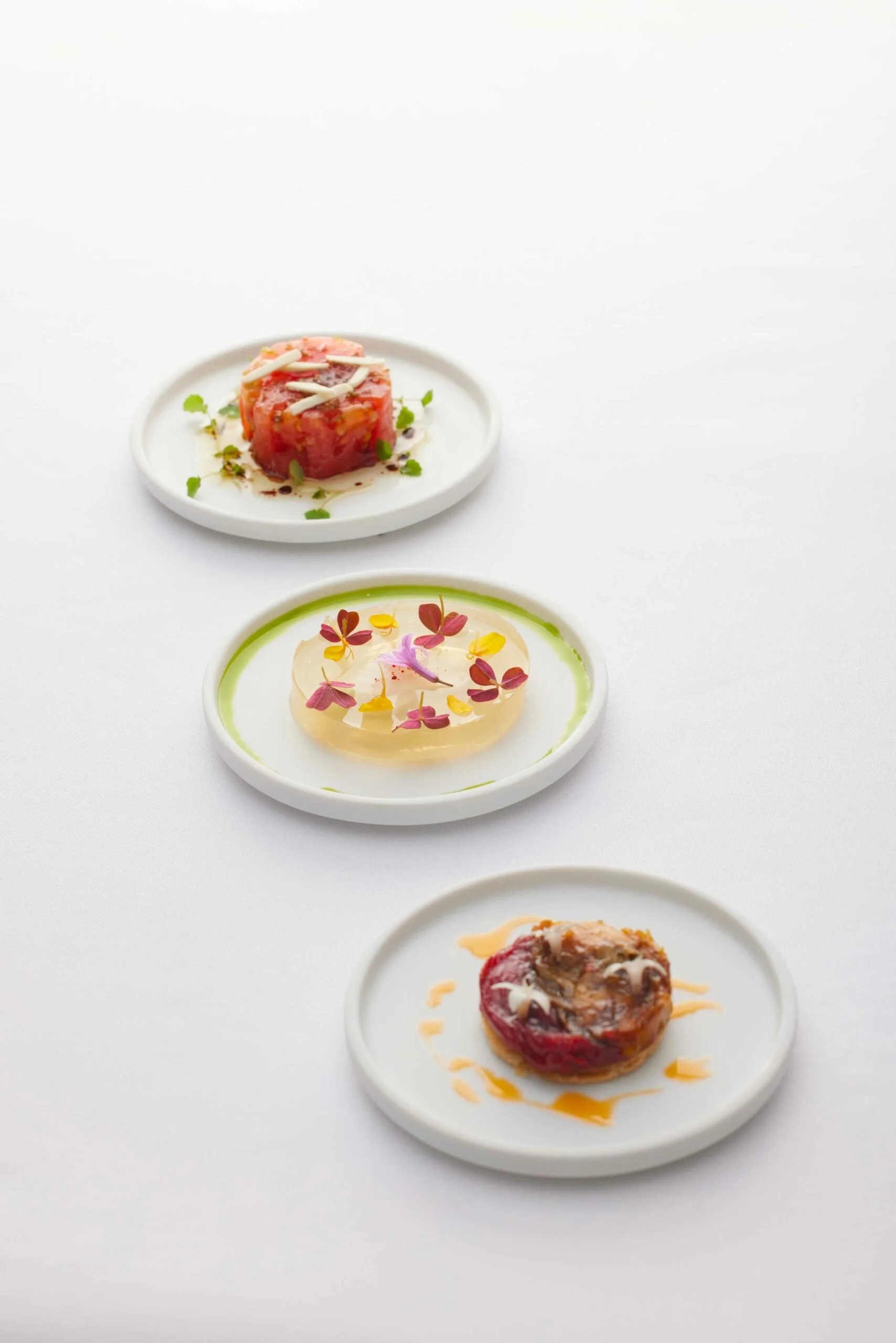 Trois petits plats de chef disposés en ligne sur une surface blanche, comprenant une entrée de tartare de viande, un plat de gelée ou aspic décoré de fleurs comestibles, et un petit plat chaud avec viande.