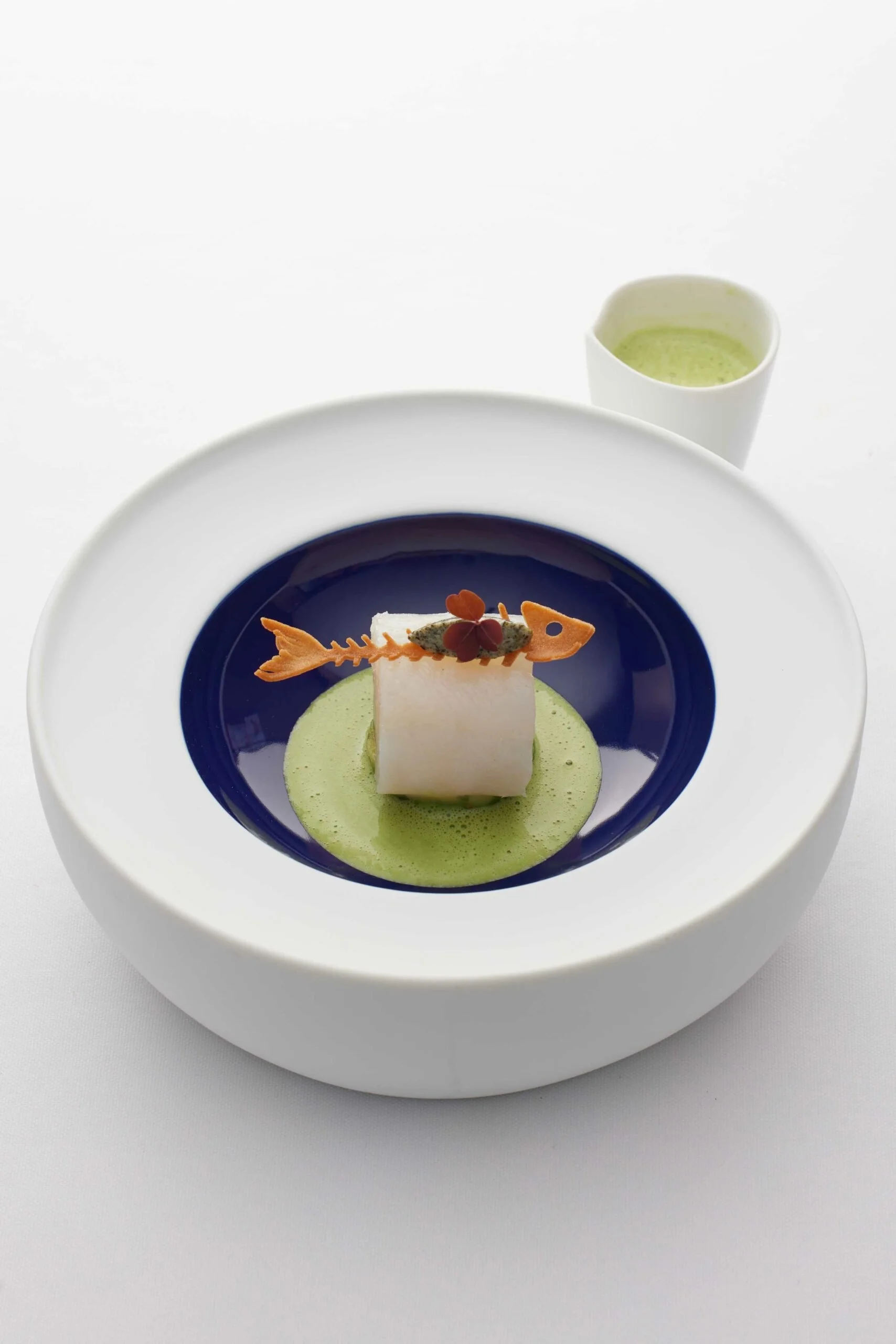 Plat gastronomique avec un morceau de poisson blanc, une sauce verte, un biscuit en forme de poisson, et une petite garniture, avec une boisson verte en arrière-plan.