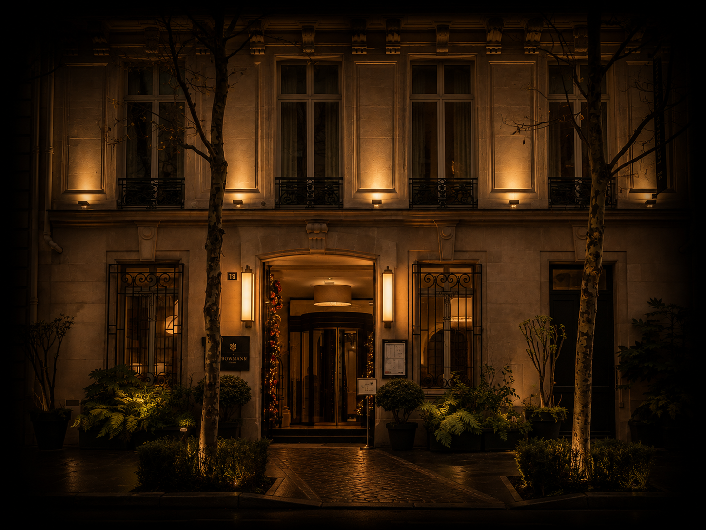 Façade d'un bâtiment élégant éclairé la nuit, avec des fenêtres et des portes ornées, des arbres devant et une décoration de Noël autour de l'entrée.