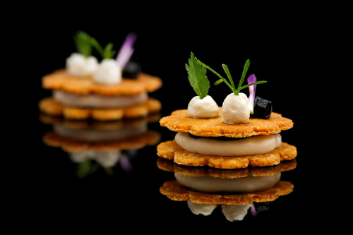 Deux amuse-gueules avec des crackers, de la crème, des herbes, et des petits morceaux de légumes, présentés sur un fond noir refletant.