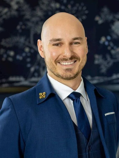 Homme souriant en uniforme bleu avec épaulette et cravate, fond flou d'une scène nocturne avec lumières et arbres.