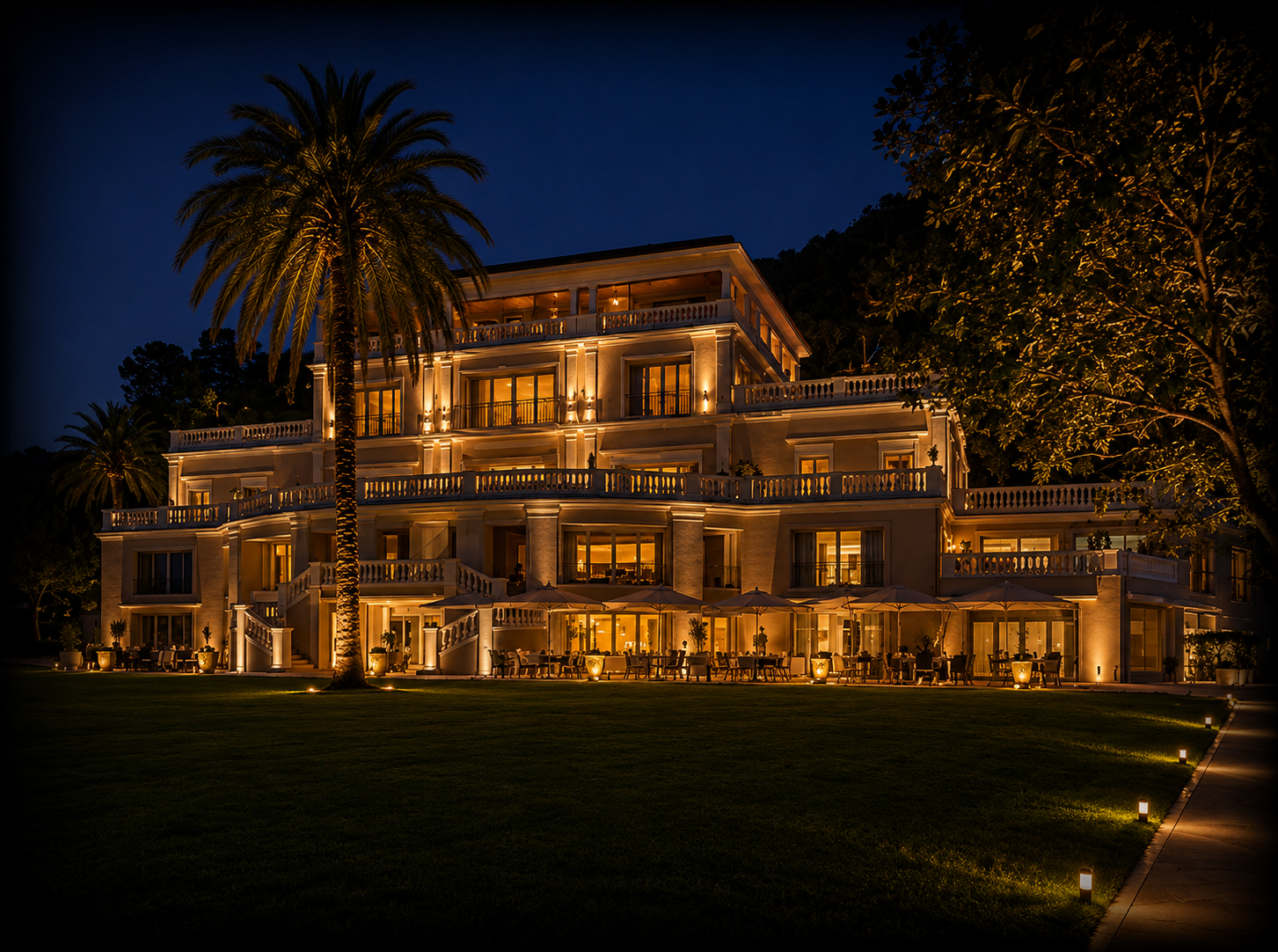Un grand hôtel illuminé la nuit avec plusieurs étages, balcons et terrasses, entouré d'arbres dont un palmier et un arbre feuillu, avec une pelouse bien entretenue et un chemin éclairé.