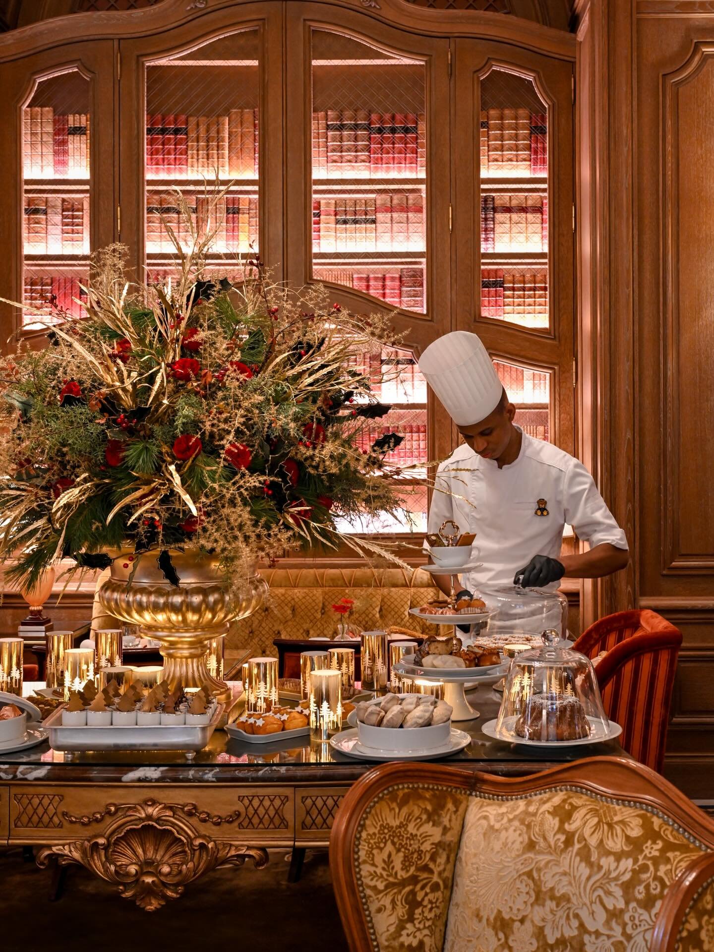 Un chef en uniforme blanc et toque préparant des desserts dans un décor élégant, avec une grande vase de fleurs, des bougies dorées, et un buffet de pâtisseries sur une table en bois ornée