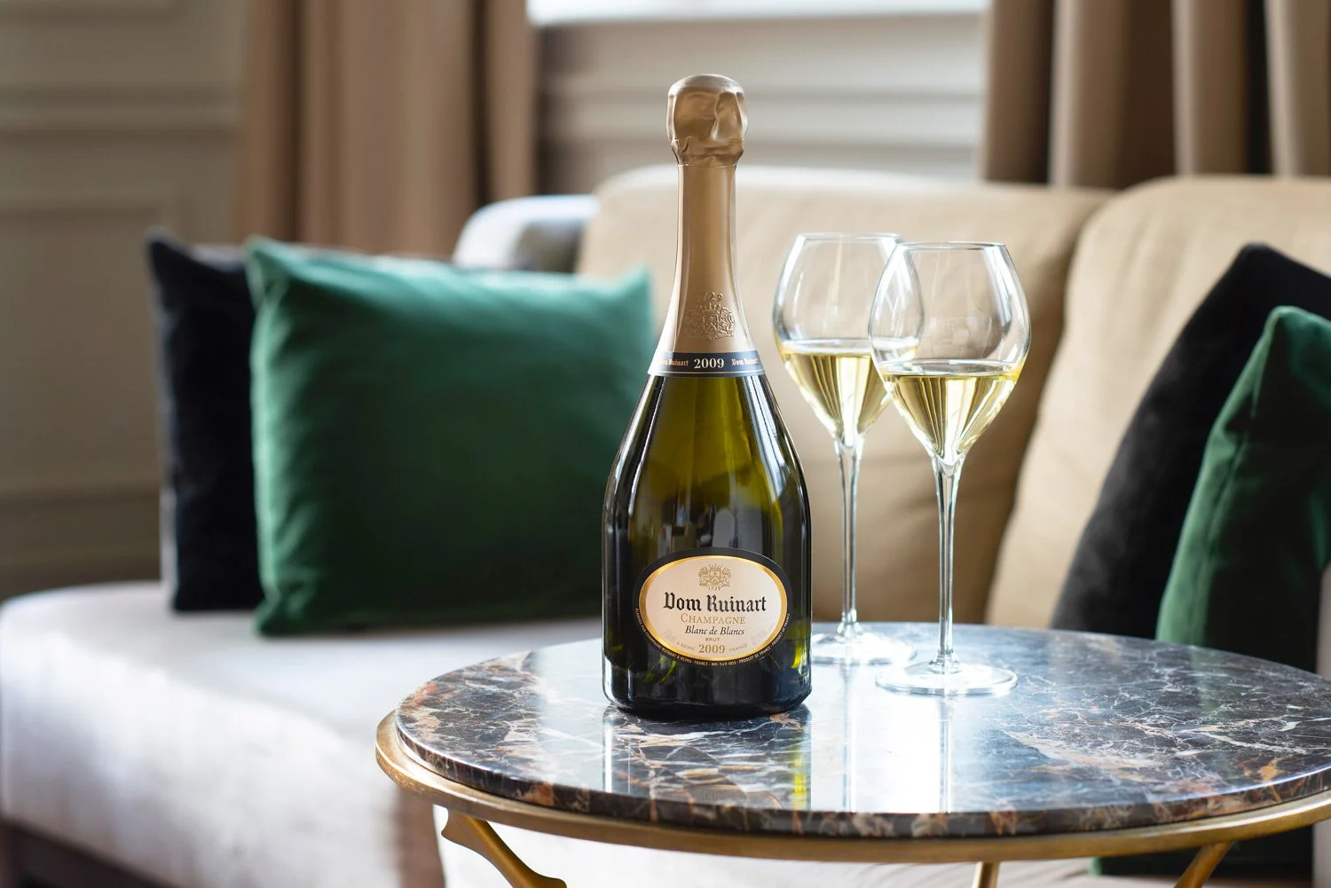 Une bouteille de champagne Dom Ruinart à côté de deux verres de vin blanc sur une table en marbre, avec un canapé et des coussins en arrière-plan.