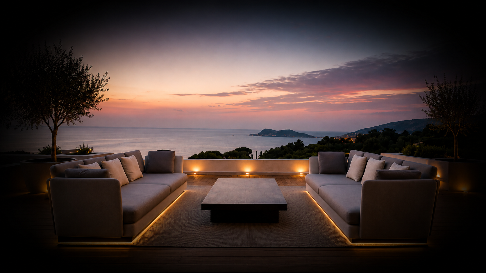 Balcon avec deux sofas modernes et une table, vue sur la mer au coucher du soleil, avec des arbres et des collines en arrière-plan.