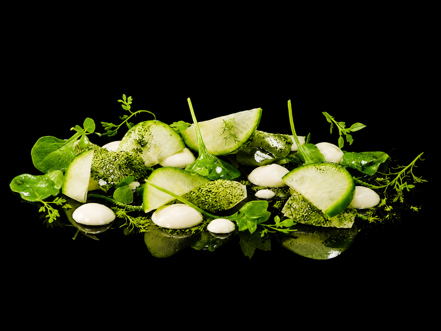 Une assiette de légumes verts, notamment des courgettes en tranches, garnie de jeunes pousses et de sauces blanches, sur fond noir.