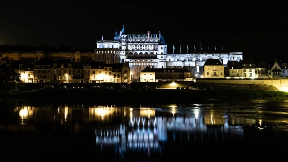 Château illuminé au bord de l'eau la nuit, avec reflection de ses lumières sur la surface.