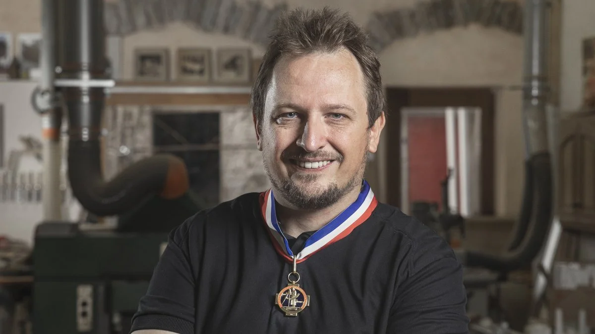 Un homme souriant portant une médaille autour du cou, dans un atelier : arrière-plan avec des machines et des meubles.
