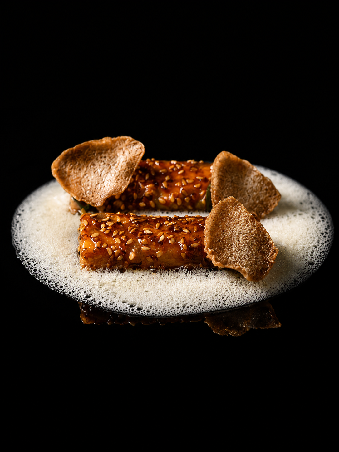 Deux rouleaux de sushi recouverts de sauce et de graines de sésame, garnis de chips croustillantes, dans de la mousse blanche, sur fond noir.