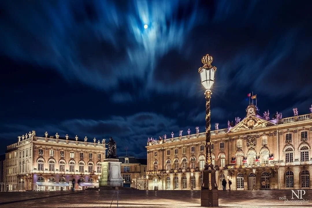 Place Royale éclairée de nuit avec un lampadaire au centre, bâtiments historiques ornés, ciel nuageux et pleine lune en arrière-plan.