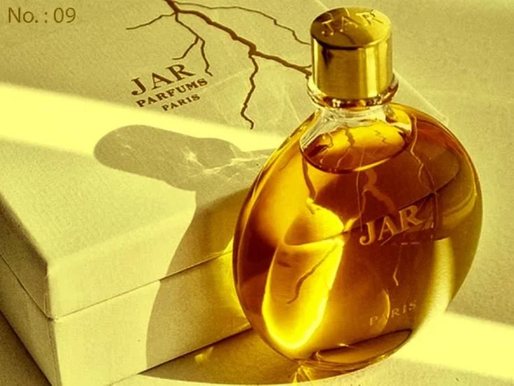 Une bouteille de parfum dorée à côté d'une boîte blanche, avec une lumière qui crée une ombre de la bouteille. La boîte est marquée "JAR PARFUMS PARIS".