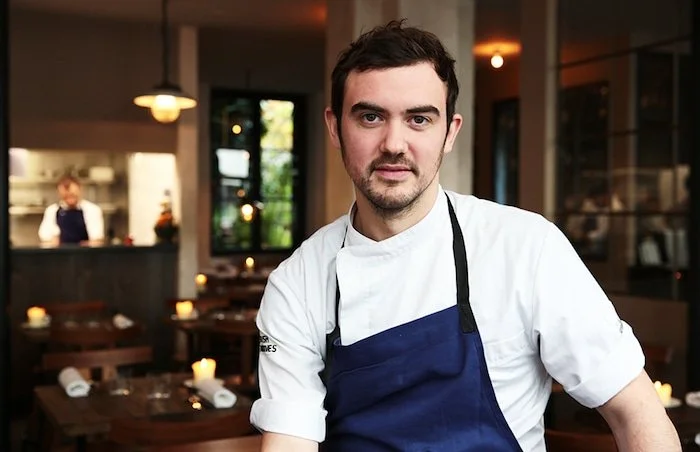 Un chef avec une veste blanche et un tablier bleu dans un restaurant, avec un décor chaleureux et une cuisinière en arrière-plan.