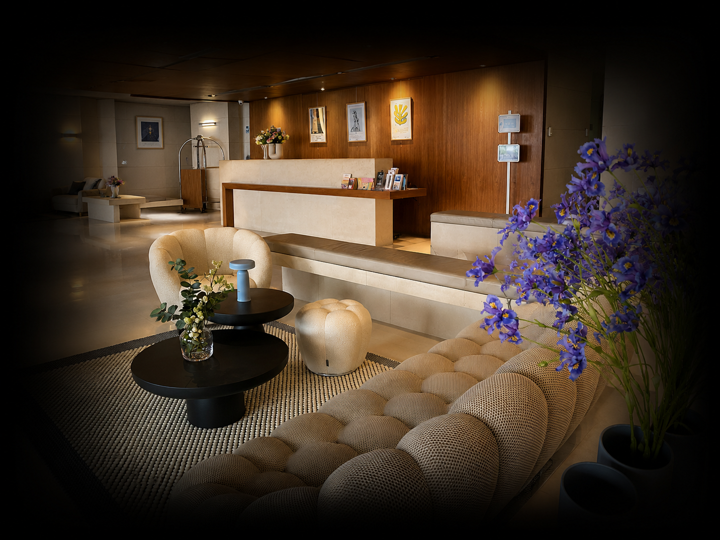 Lobby d'hôtel élégante avec canapé beige, fauteuil, table basse noire avec vase de fleurs, mur en bois avec œuvres d'art, réception en arrière-plan, et fleurs violettes dans un pot