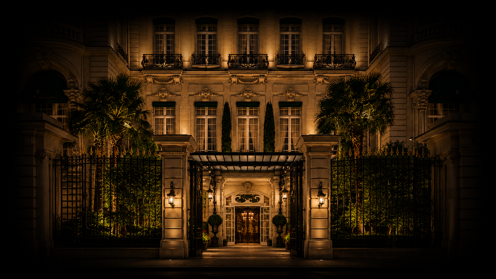 Une grande maison élégante éclairée la nuit, entourée d'une grille en fer forgé et de palmiers, avec une entrée illuminée.