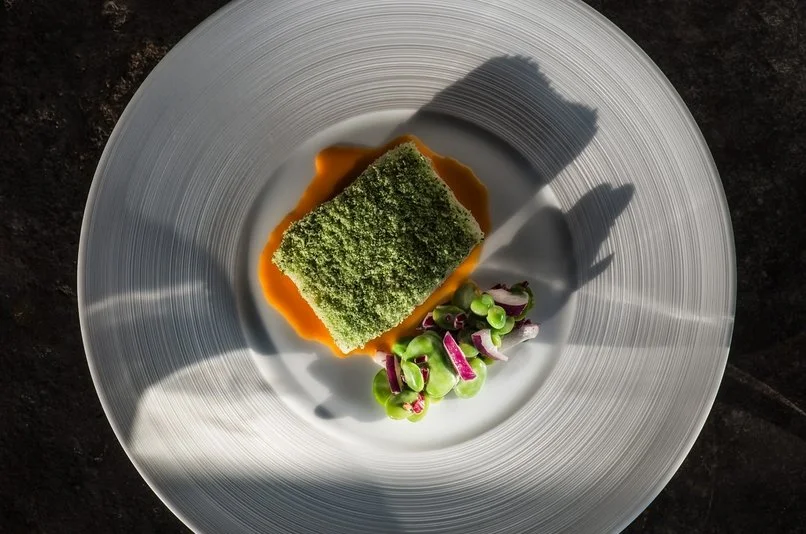 Plat gastronomique avec un morceau rectangulaire recouvert de croustillant vert, nappé de sauce orangée, accompagné d'une garniture de pois verts et radis coupés en rondelles, servi dans une assiette blanche
