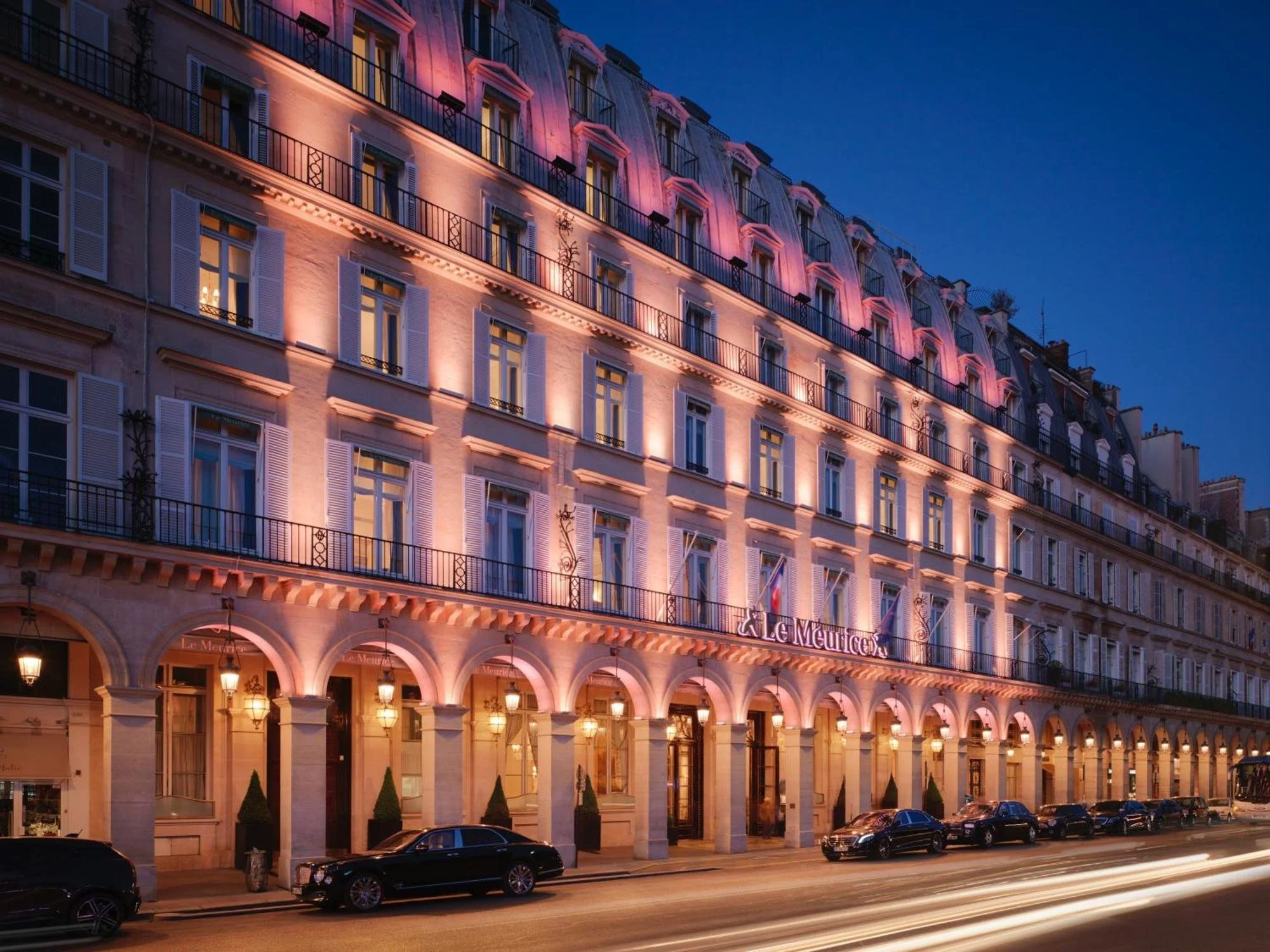 Bâtiment élégant avec de grandes fenêtres et des balcons, illuminé en rose et violet, situé sur une rue parisienne la nuit