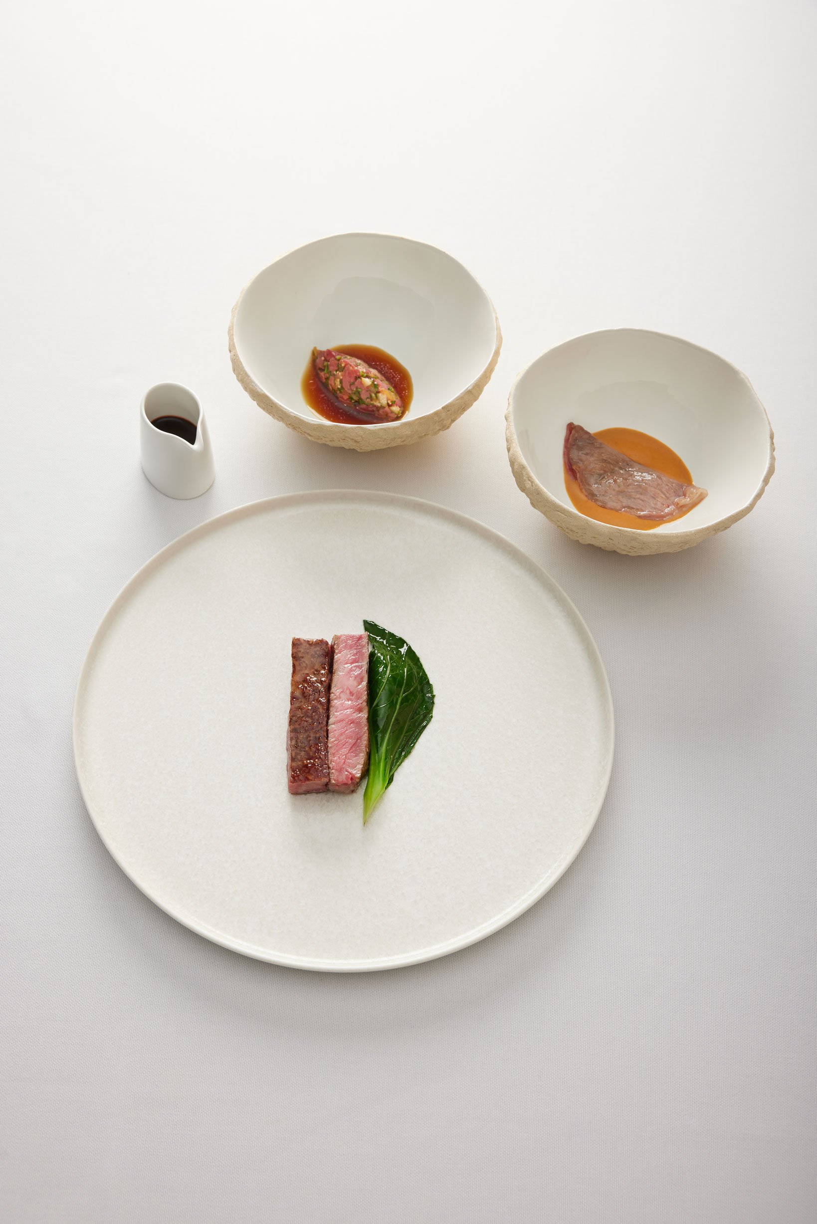 Assiette avec trois morceaux de viande et une feuille de légumes vert, deux bols avec sauces ou purées, petite cruche avec sauce noire, présentation minimaliste sur une nappe blanche.