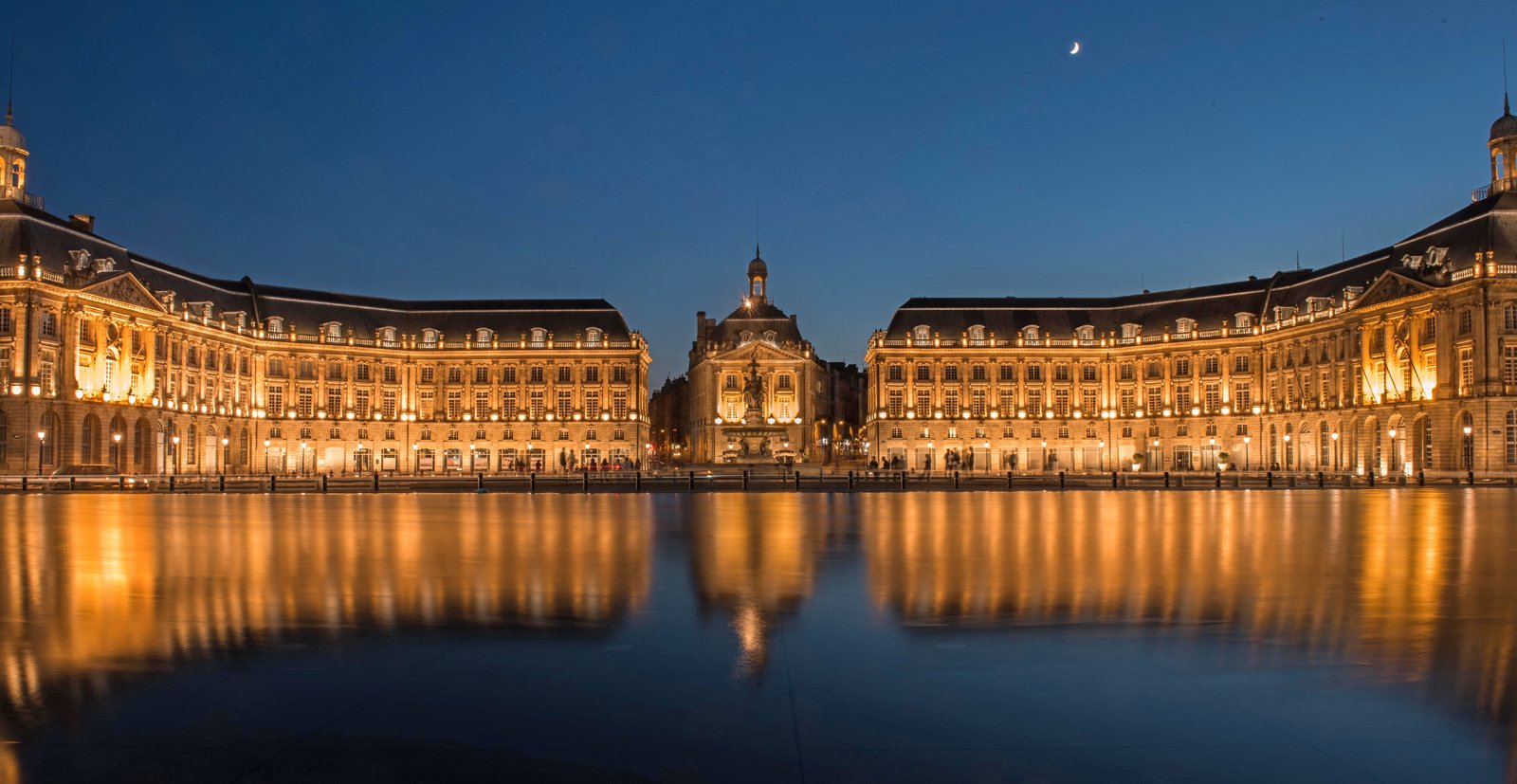Vue nocturne d'une grande place historique avec des bâtiments illuminés de chaque côté, un ciel clair avec une lune croissante, et un reflet dans l'eau au premier plan.