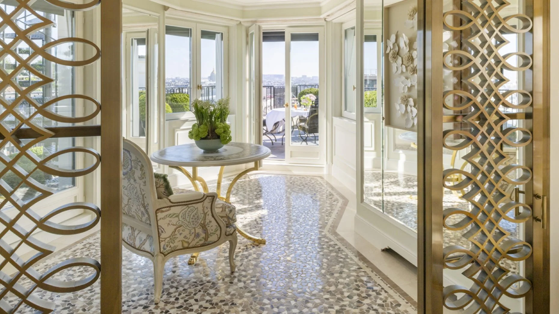 Salle à manger lumineuse avec balcon, table rond, fauteuils en tissu brodé, plantes vertes en vase, et vue sur Paris