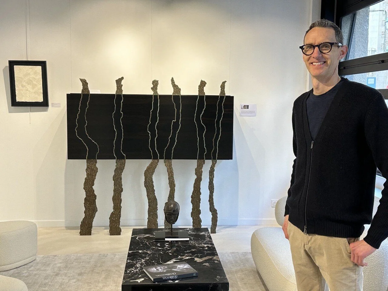 Un homme souriant portant des lunettes noires, chemise noir et pantalon beige, dans une galerie d'art avec plusieurs œuvres et sculptures modernes, y compris une sculpture de tête noire sur une table en marbre noir, et des œuvres murales en arrière-plan.