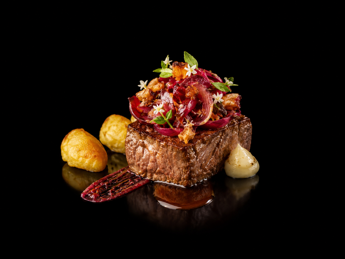 Un plat de viande de bœuf grillée, garnie d'oignons rouges, de microgreens et de fleurs comestibles, avec de petites pommes de terre ou gnocchis et une sauce ou purée blanche, présenté sur un fond noir.