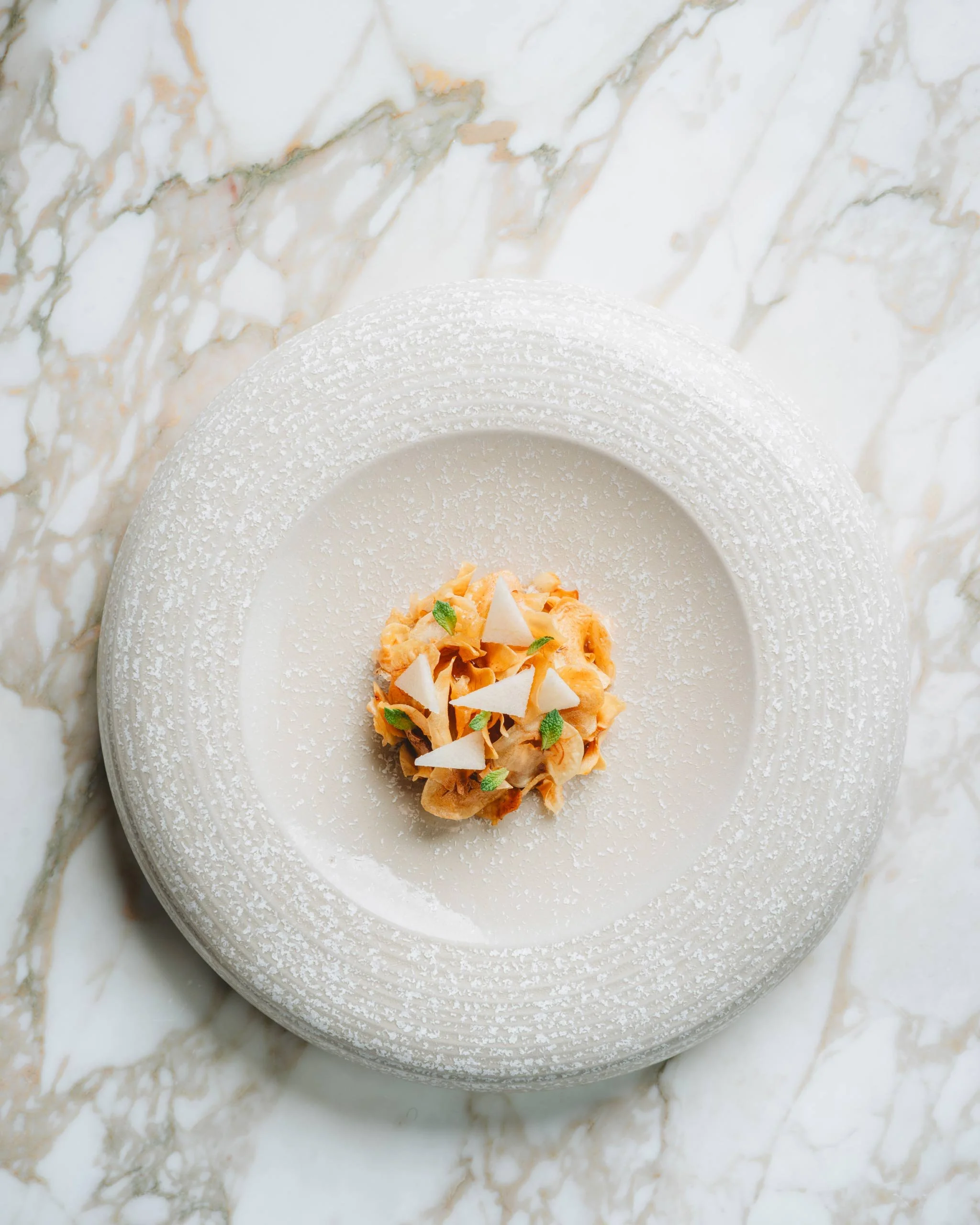Plat de dessert avec un petit gâteau décoré de morceaux de blanc et de feuilles vertes, présenté sur une assiette blanche texturée, posé sur une surface en marbre blanc veiné.