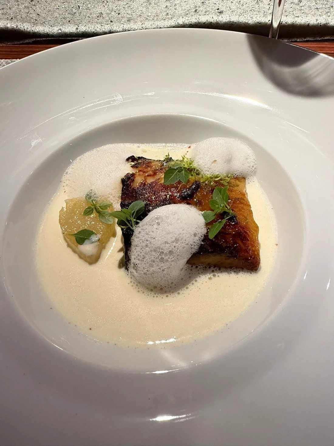 Plat de restaurant avec un morceau de poisson ou de viande, mousse ou sauce blanche, garniture de feuilles vertes, citron, et une mousse blanche en surface, servi dans une assiette blanche