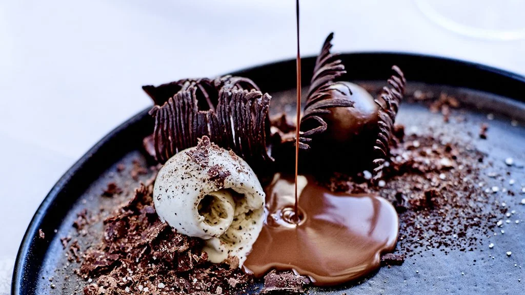 Un gâteau au chocolat décoré de morceaux de chocolat, de pâte à sucre blanche et d'une boule en forme de tête, avec du chocolat fondu versé dessus.
