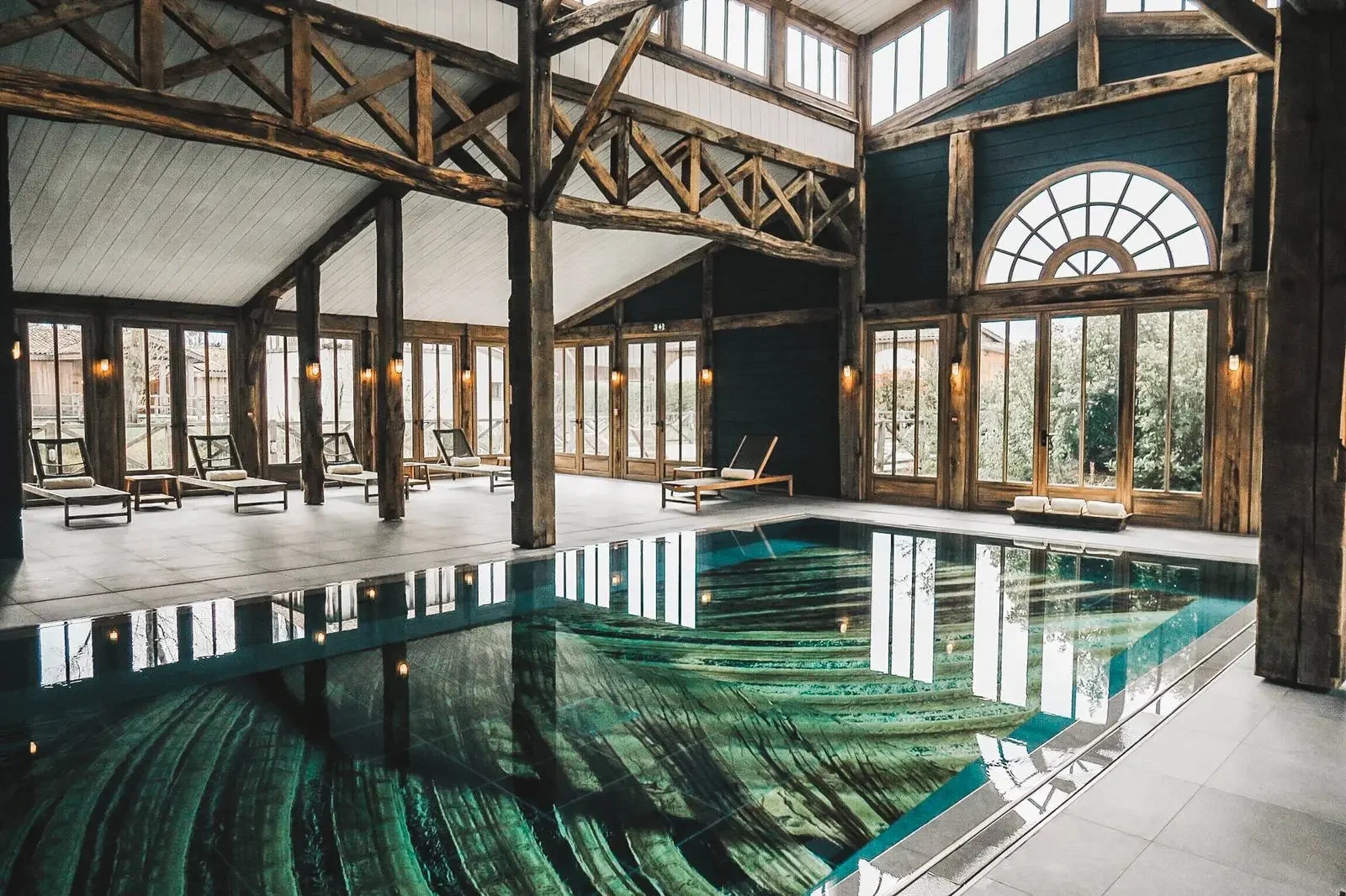 Intérieur d'une piscine dans une salle en bois avec de grandes fenêtres, des chaises longues en bois et un style rustique.