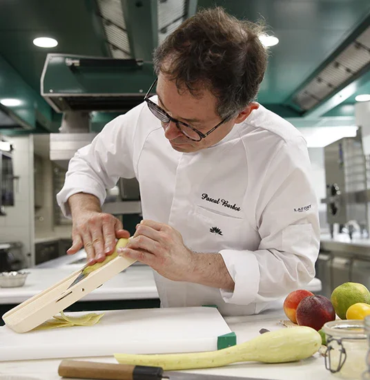 Chef professionnel dans une cuisine de restaurant, portant un tablier marron et une chemise blanche, se tenant derrière un plan de travail blanc avec des figues et un gâteau rond, tout en ajustant ses lunettes et regardant vers la caméra.