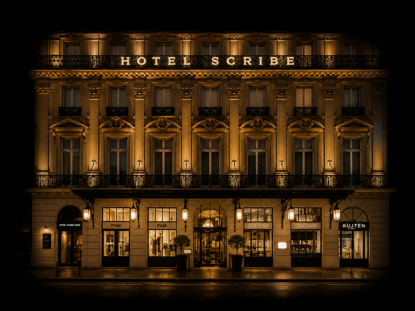 Immeuble élégant éclairé la nuit avec le panneau 'HOTEL SCRIBE' en haut, façades ornées, boutiques au rez-de-chaussée.
