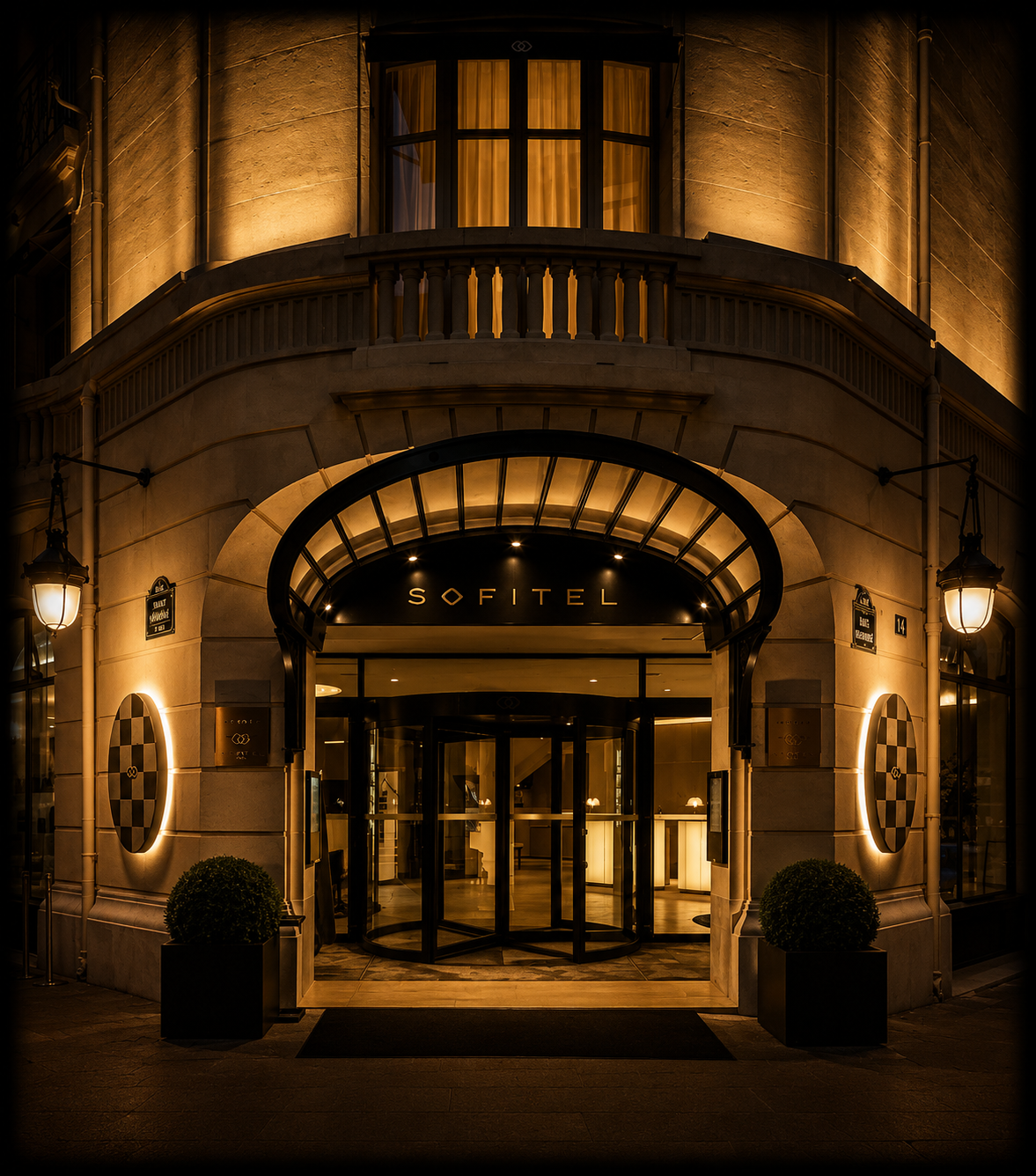 Entrée d'un hôtel Sofitel illuminé la nuit, avec deux lampes extérieures, des plantes dans des pots noirs, et un arc en verre autour de l'entrée.